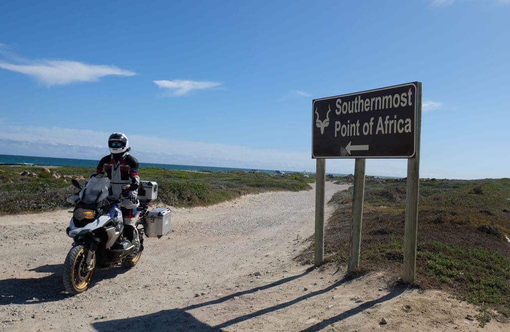 One of the bikers sits on their bike next to a sign saying 'Southernmost point of Africa'.